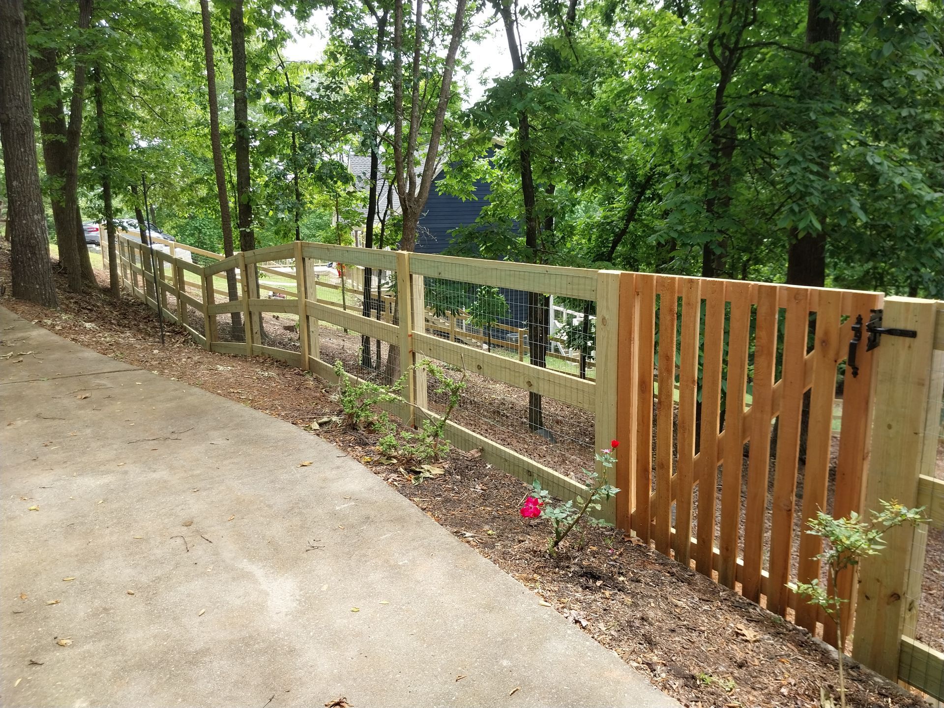 Wooden fence along a sloped wooded path with a gate and concrete driveway edge