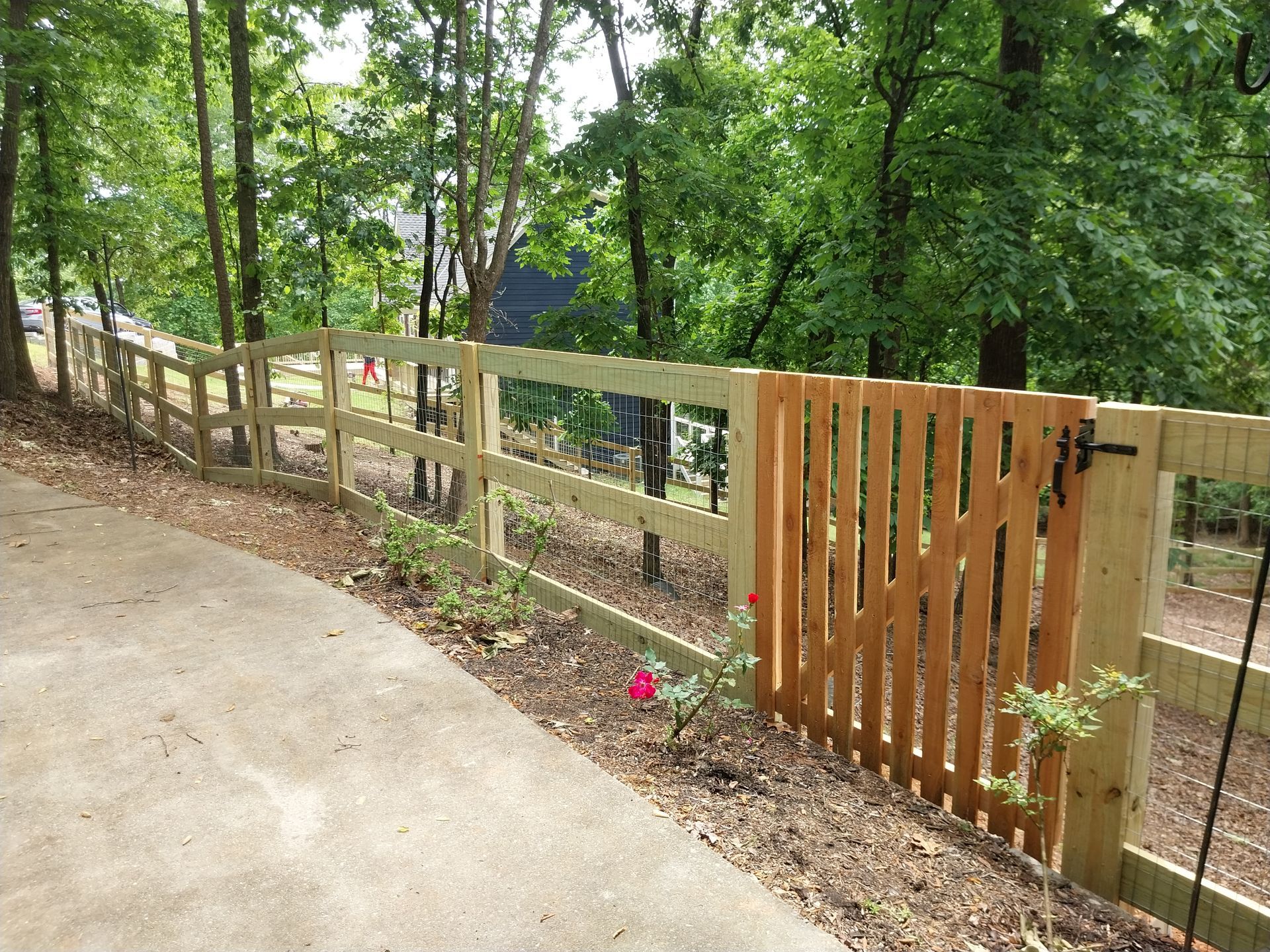 Wooden fence along a wooded path beside a concrete walkway, with a gate and orange flowers.