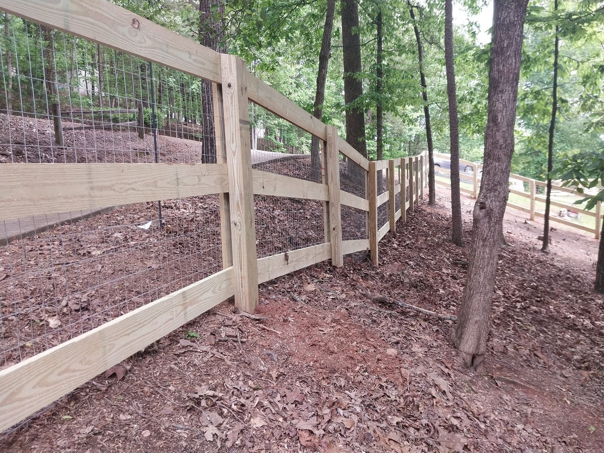 Wooden split-rail fence along a wooded dirt path on a sloped hillside.