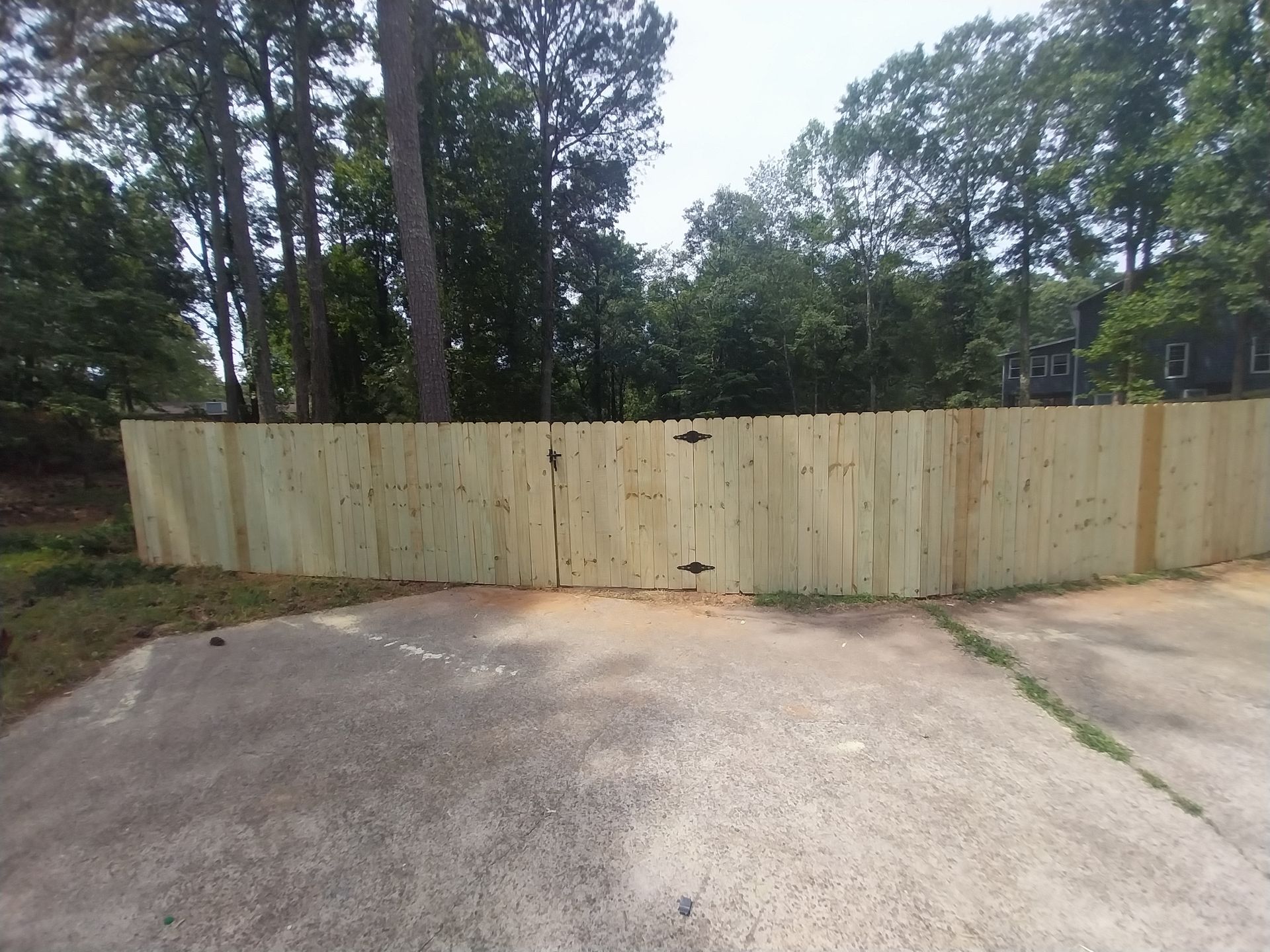 Concrete driveway facing a light wooden fence with trees and a wooded yard behind it