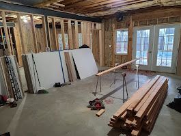 Interior renovation in progress, framing, drywall, lumber, and tools scattered on the concrete floor. French doors visible.