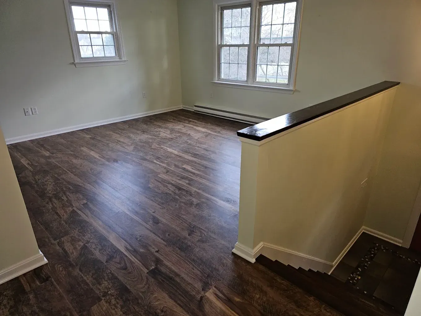 Empty living room with dark wood floors, two windows, and a stair railing on the right.