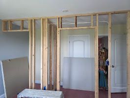 Interior view of a room under construction; wooden studs form the frame of walls and doorway, with sheetrock panels present.