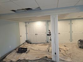 Basement undergoing renovation, with drop cloths, paint supplies, and damaged ceiling. White doors and column.