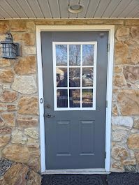Gray door with white trim set in stone wall. Includes glass panel, light fixture, and doormat.