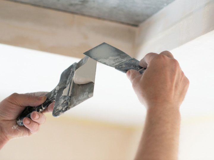 Hands using two putty knives to smooth drywall compound on a ceiling corner.