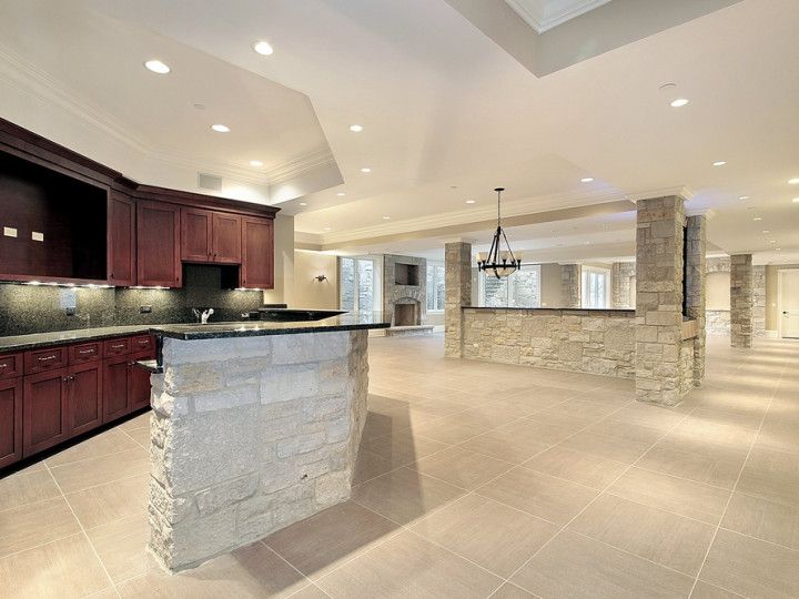 A large, open basement with a stone bar and dark wood cabinets. Beige tiled floors and recessed lighting.
