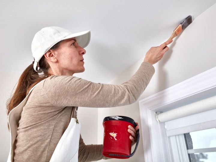 Woman in white cap painting a ceiling, holding a red paint bucket, near a window.