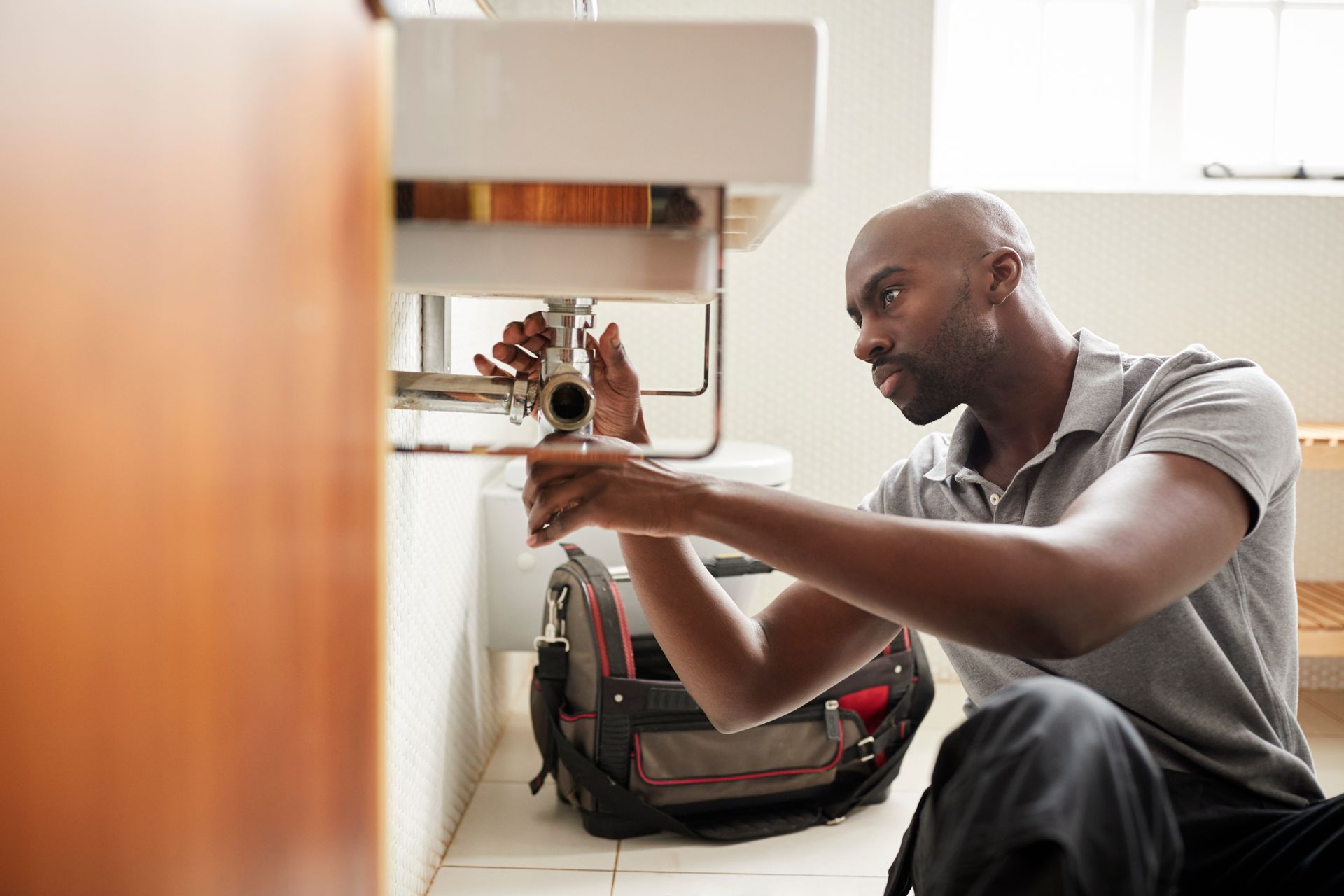 A plumber crouched under a bathroom sink, inspecting the pipe fittings with a tool bag beside him