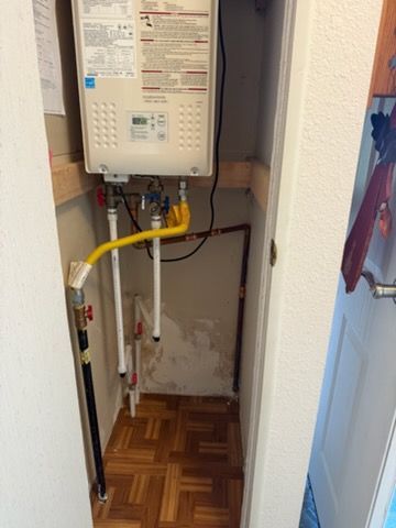 Wall-mounted gas water heater in a narrow closet with exposed pipes and wooden floor.