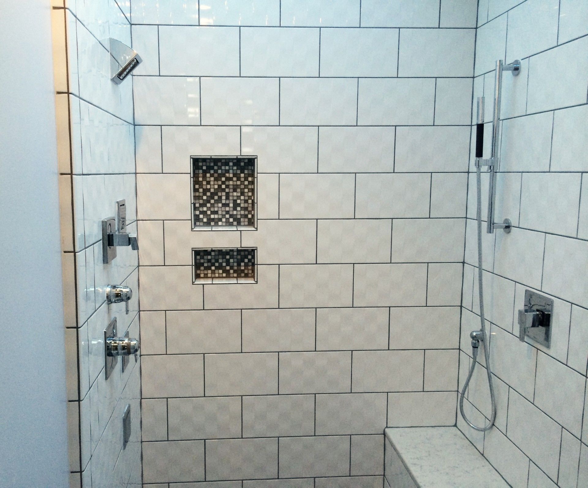 Tiled shower with shower head, handheld sprayer, and built-in shelves. Gray and white tiles.