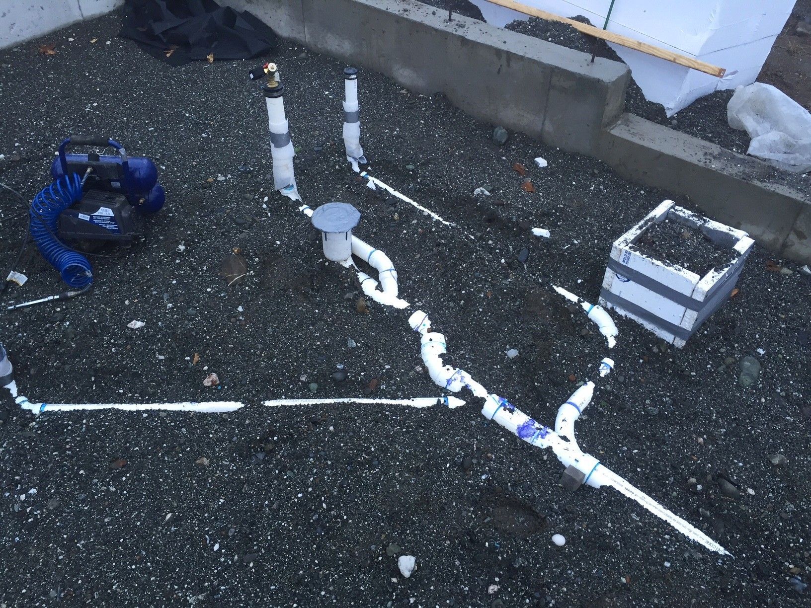 Pipes and drainage system installed on gravel ground, near a concrete wall and a small box.