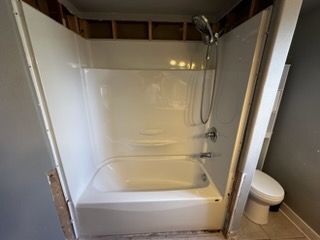 White alcove bathtub and shower unit in a bathroom with partially exposed framing. Toilet to the right.