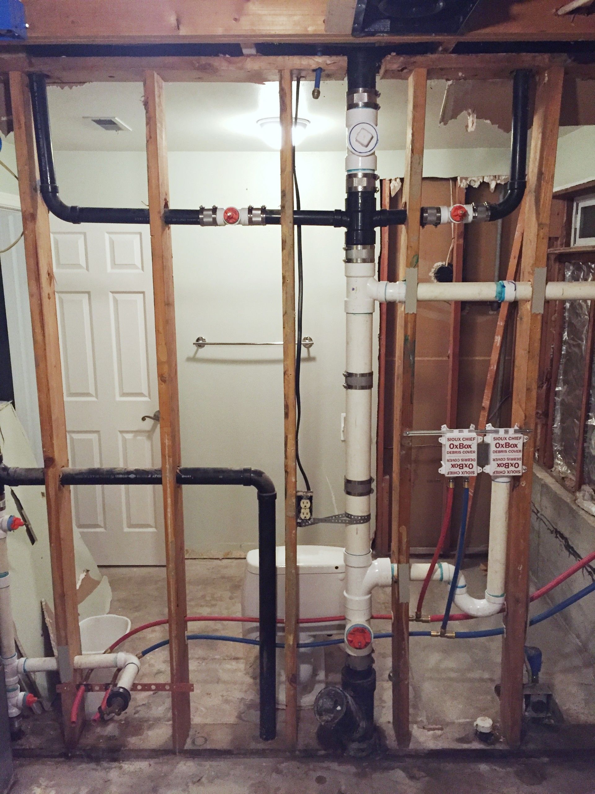 Plumbing pipes and framing in a bathroom under construction; white and black pipes, red and blue water lines.