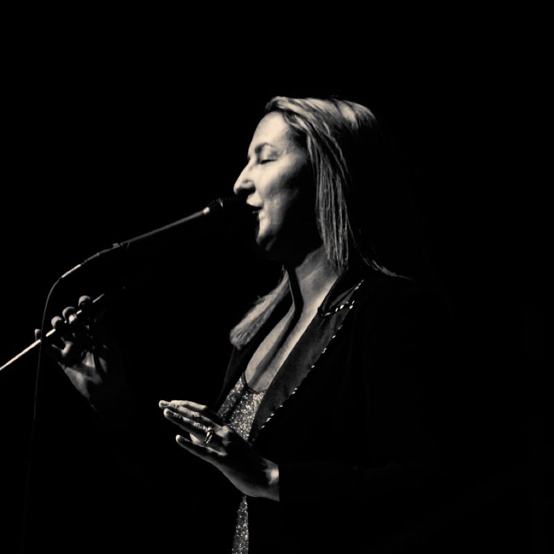 Elizabeth Jones performing as Christine McVie with Tell Me Lies - Fleetwood Mac Tribute Woman singing into a microphone on stage, in a dark setting, with arms gesturing.