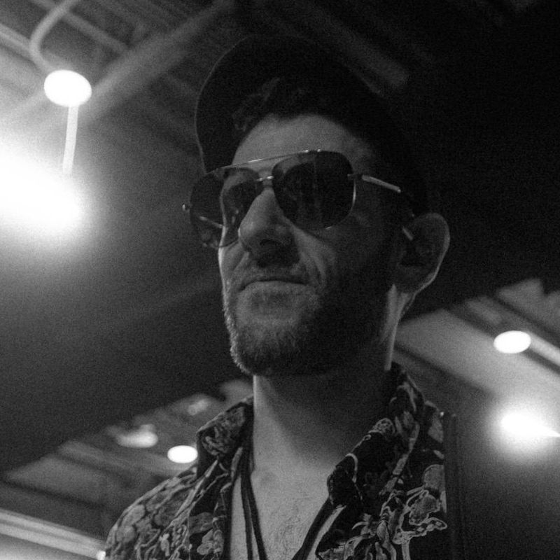 Chris Monteleone performing as John McVie with Tell Me Lies - Fleetwood Mac Tribute Man with sunglasses and hat, looking up, in a dimly lit room, wearing a patterned shirt.