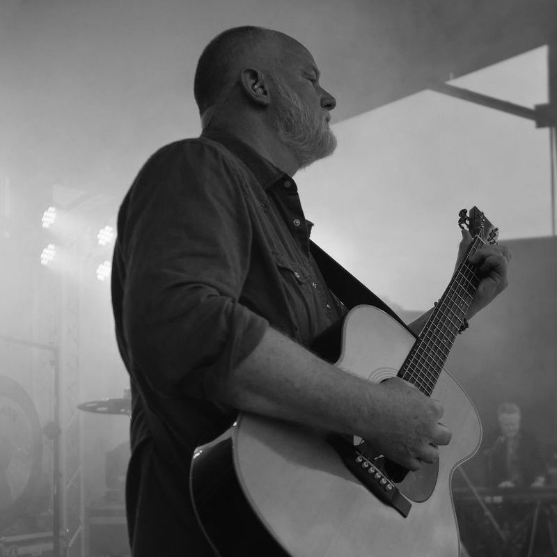 Jeff Johnson performing with Tell Me Lies - Fleetwood Mac Tribute Man with a gray beard playing an acoustic guitar on a stage, bathed in stage lighting.