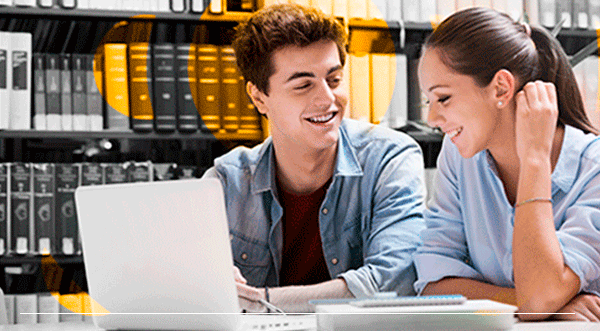 Un hombre joven y una mujer sonriendo ante una computadora portátil en una biblioteca; libros en el fondo.
