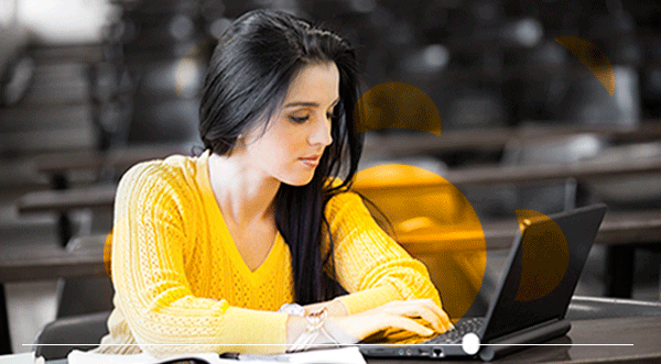 Mujer con suéter amarillo escribiendo en una computadora portátil en un aula, concentrada.