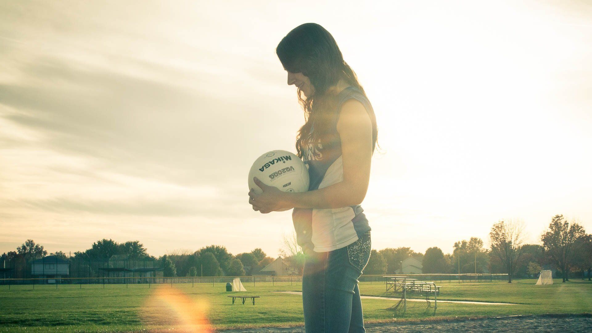 High School senior girl holding a volleyball on a sunlit field at sunset