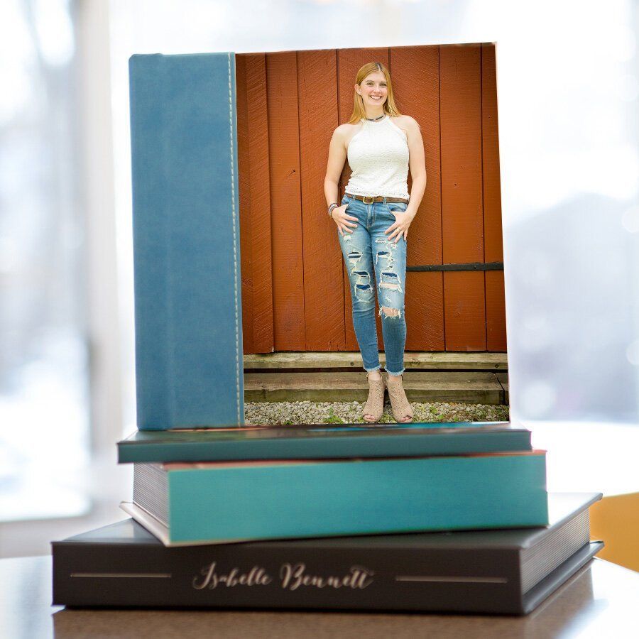 A custom designed High School Senior photo album on a book stack of a girl in a white top and ripped jeans against a red wall