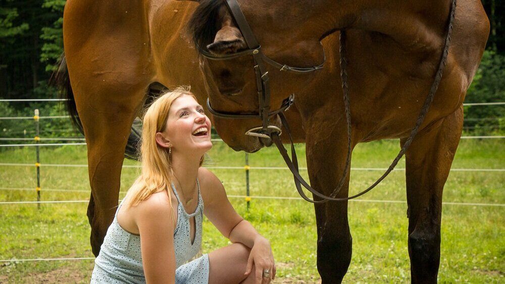 High school senior girl in Fishers crouching and smiling beside a brown horse in a grassy fenced field
