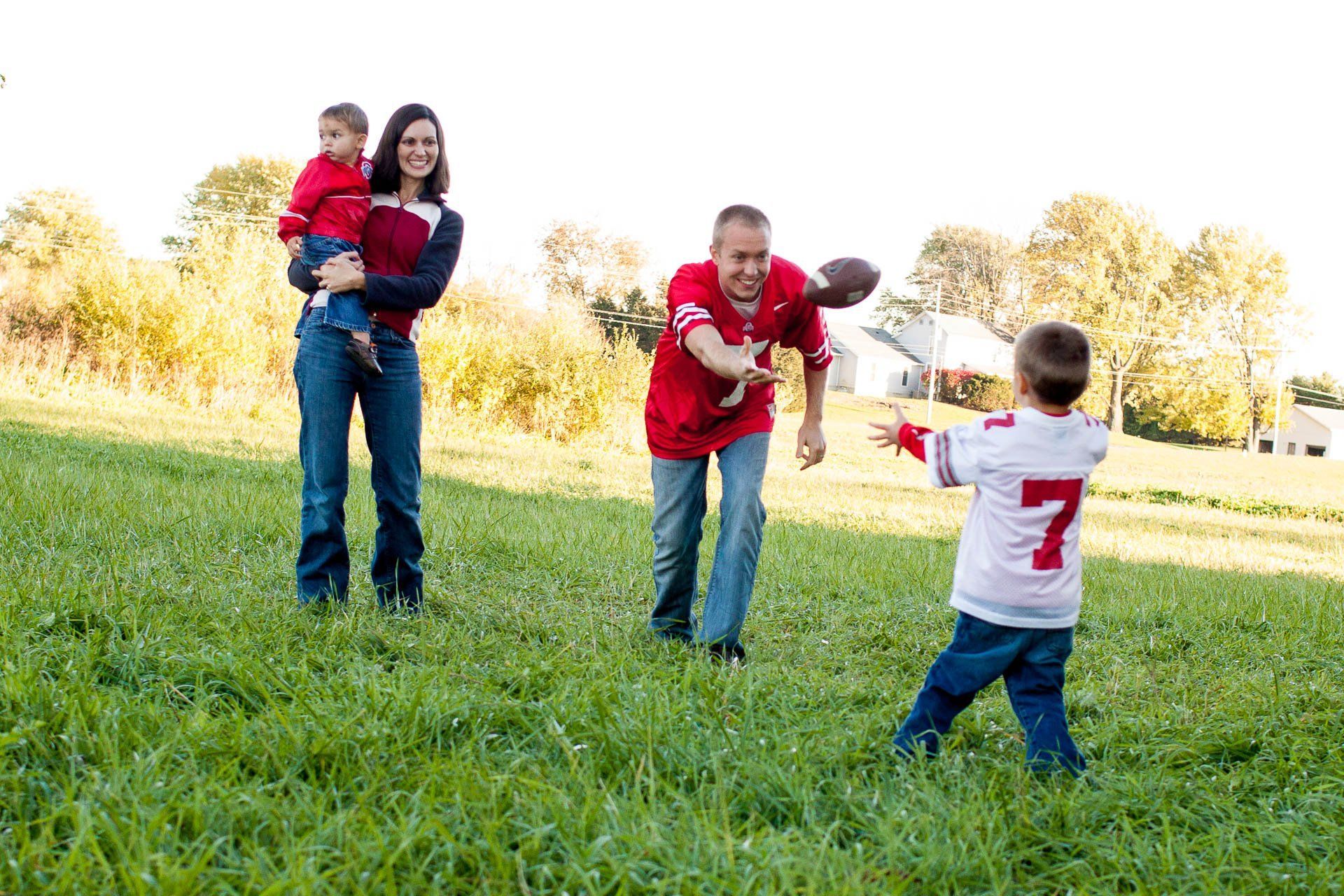 Family with two young boys playing catch in a sunny grassy park in Fishers, IN while wearing sports jerseys.