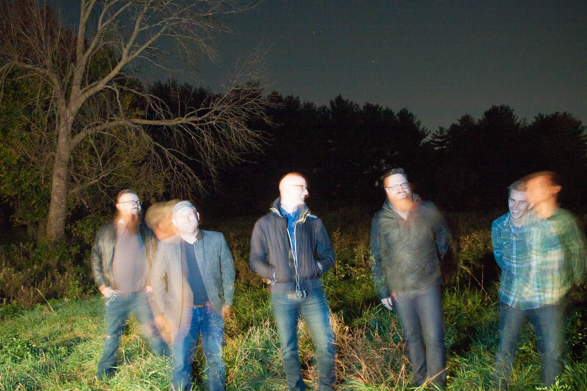 Five people standing in a grassy field at night, softly lit and slightly blurred.