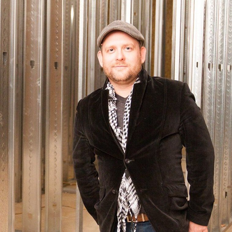 Headshot in Fishers Indiana of a man standing in front of a metallic wall, wearing a cap, black blazer, and patterned shirt.