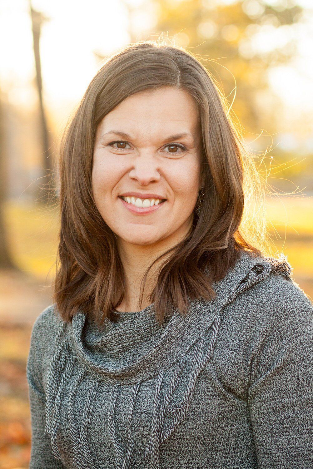 Headshot of a female Noblesville Indiana business owner smiling outdoors in a gray sweater with a soft, sunlit background