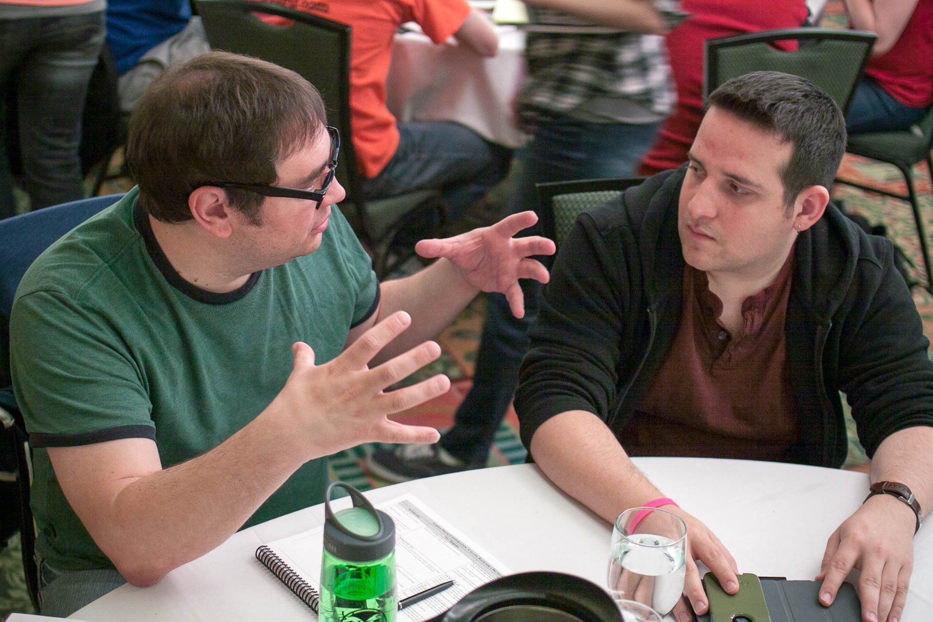 Two men talking at a table, one gesturing animatedly during a meeting or discussion.