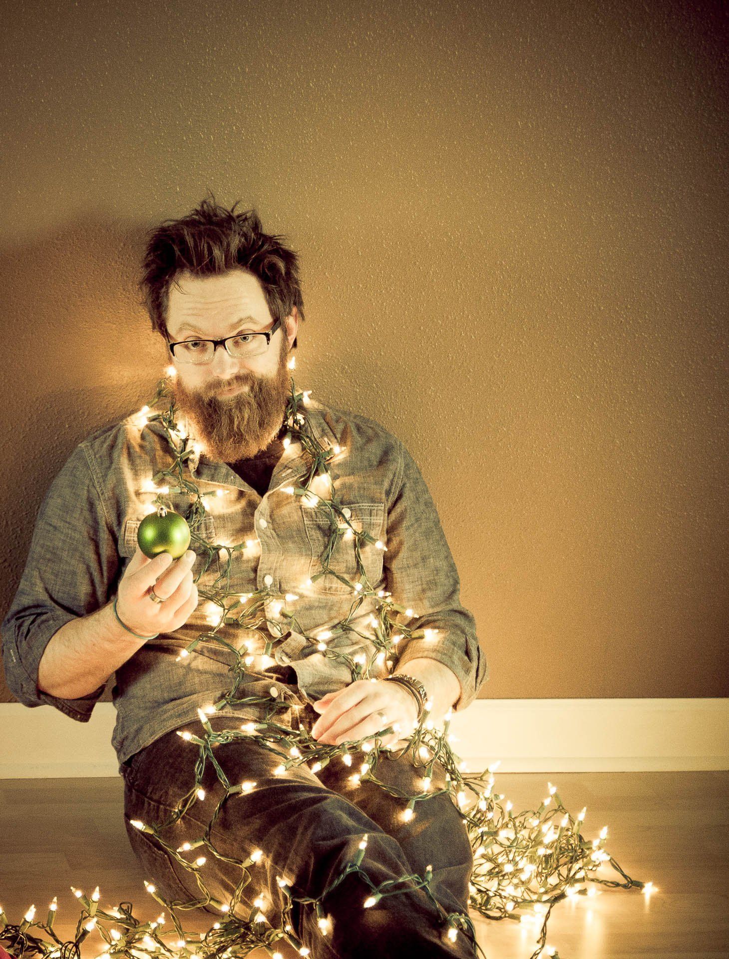 Designer sitting on the floor tangled in glowing string lights, holding a small green ornament against a plain wall.