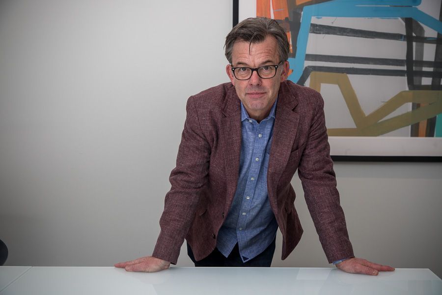 Man in glasses leaning on a white table in an office, wearing a maroon blazer and blue shirt.
