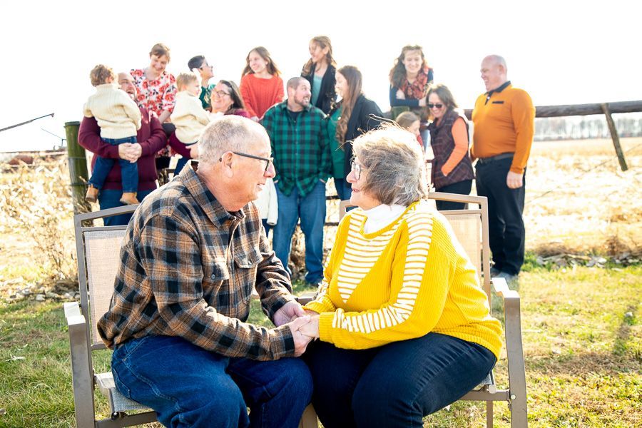 An elderly couple sits facing each other outdoors while their extended family stands behind them in a sunny field in Fishers.