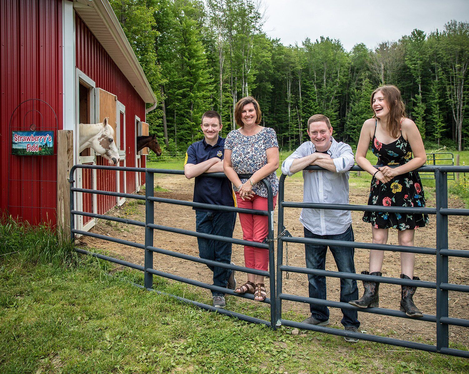 Family of four smiling by a metal gate outside a red barn with horses in a green rural setting in Westfield, IN.
