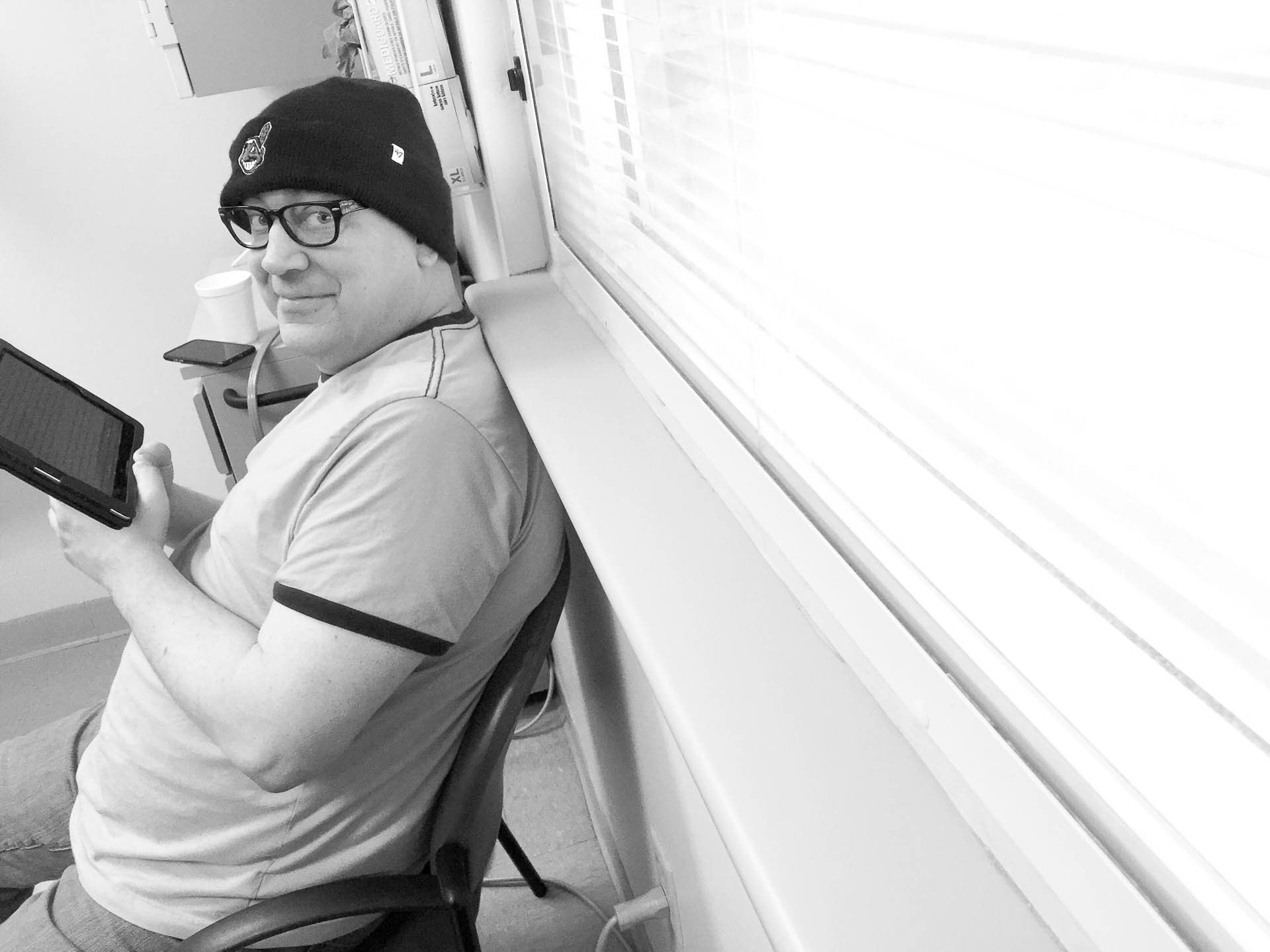 Consultant in a beanie and glasses smiling while holding a phone, sitting beside a window indoors.