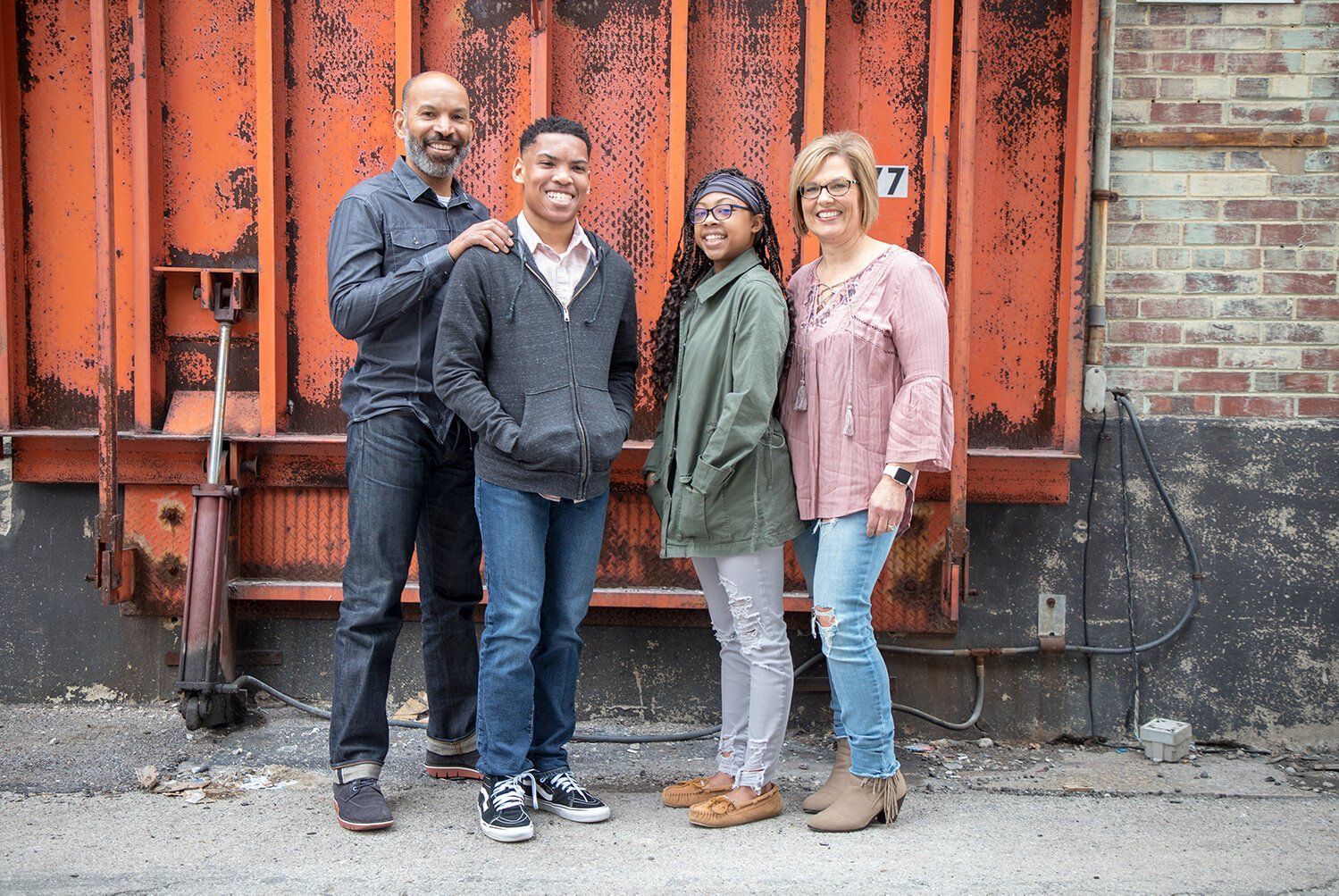 Family of four people stand smiling in front of an orange industrial wall and brick building in Zionsville.