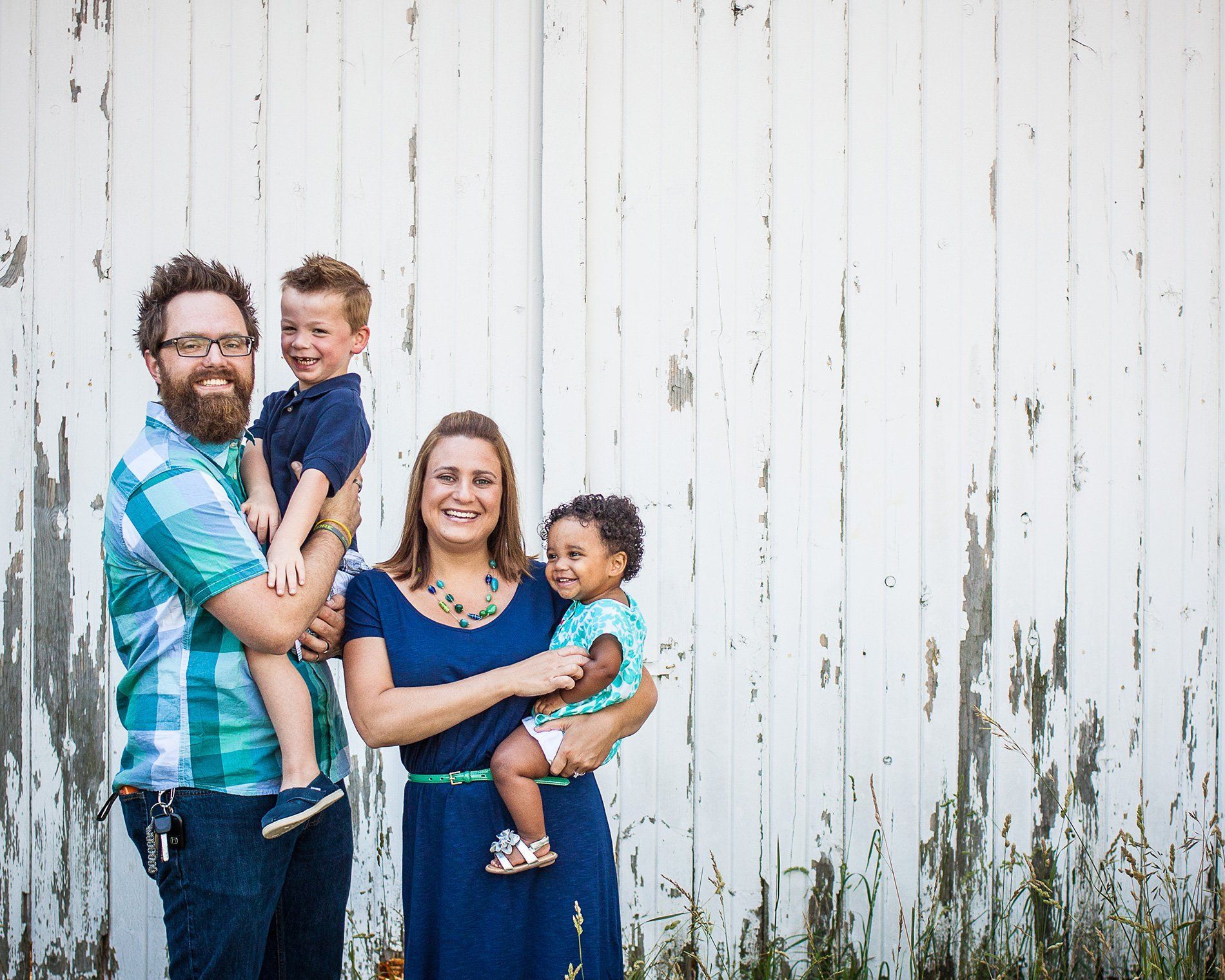 Family of four smiling and holding two children in front of a weathered white wooden wall in Fishers, Indiana