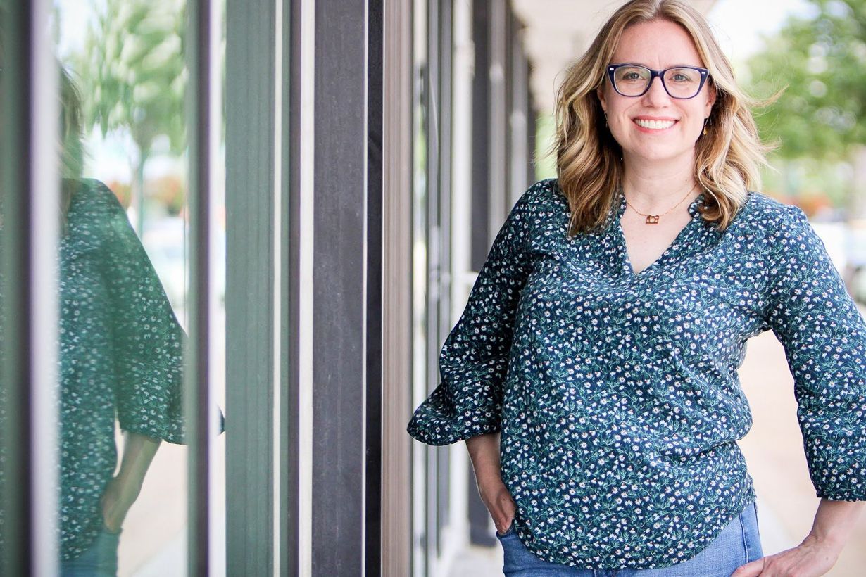 Owner Kristin Madeja leaning by a reflective window, smiling in a blue patterned blouse.