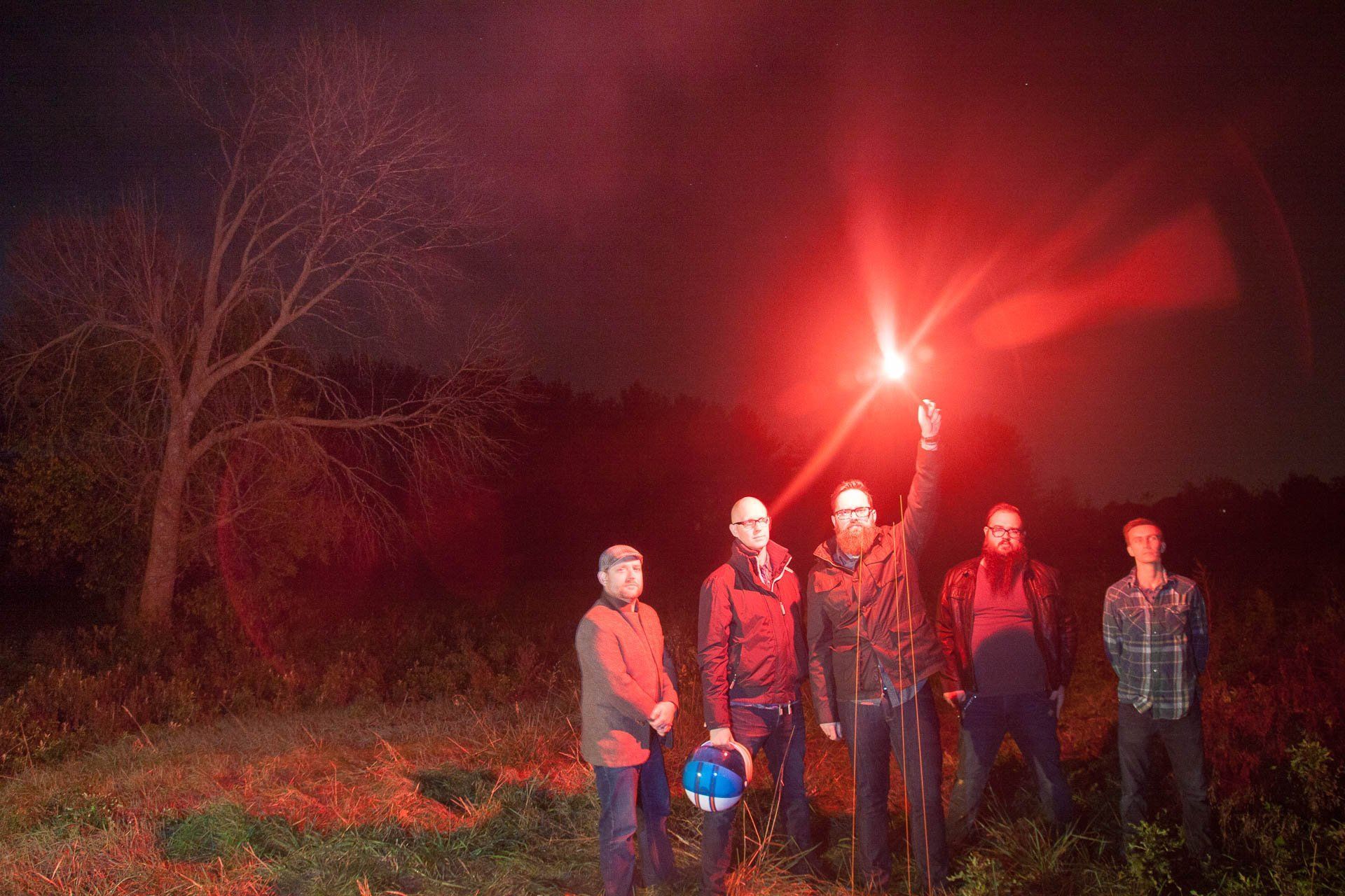 Five musicians standing outdoors at night, one holding a bright red flare.