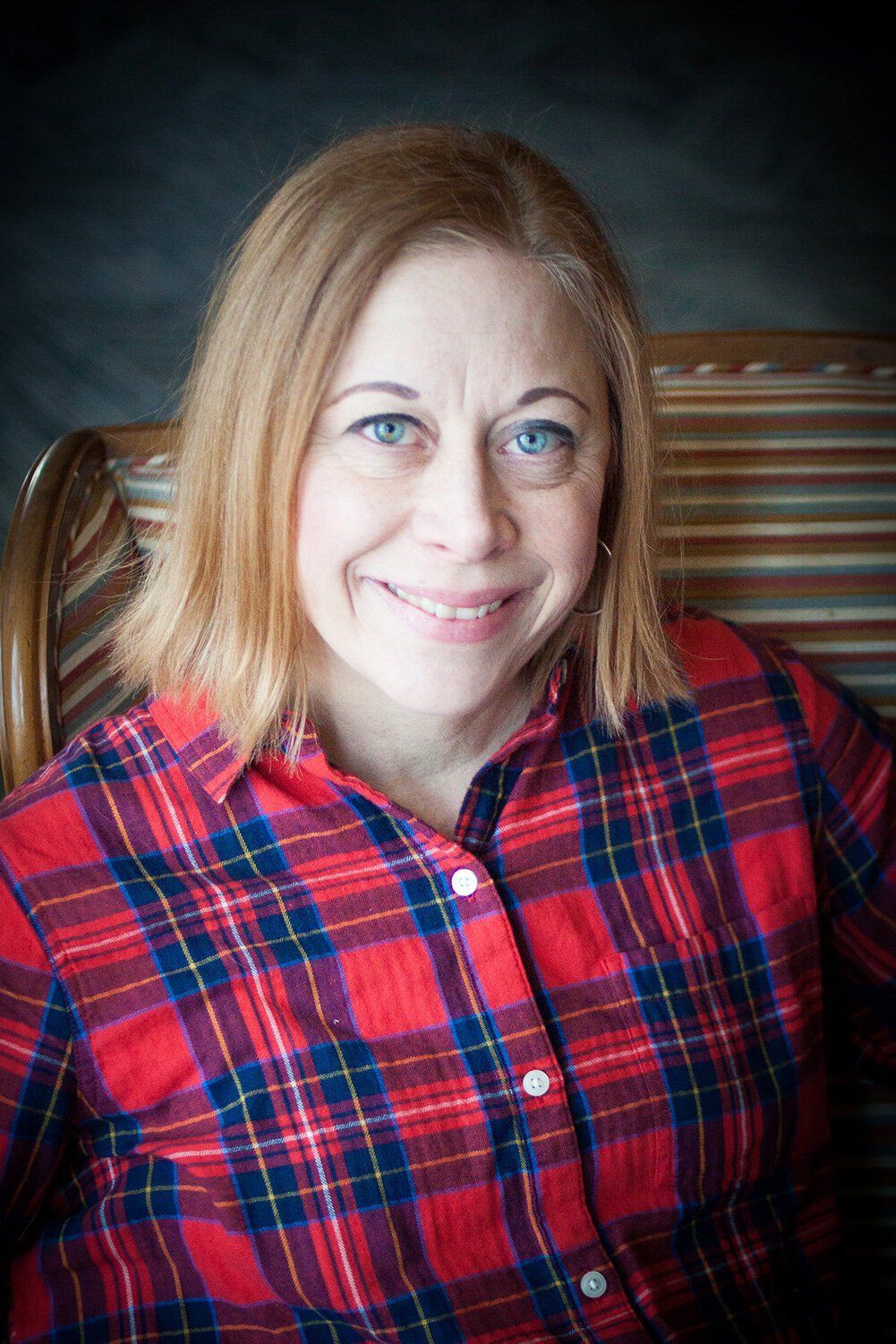 Headshot of an Carmel woman smiling in a red plaid shirt, seated against a dark background.