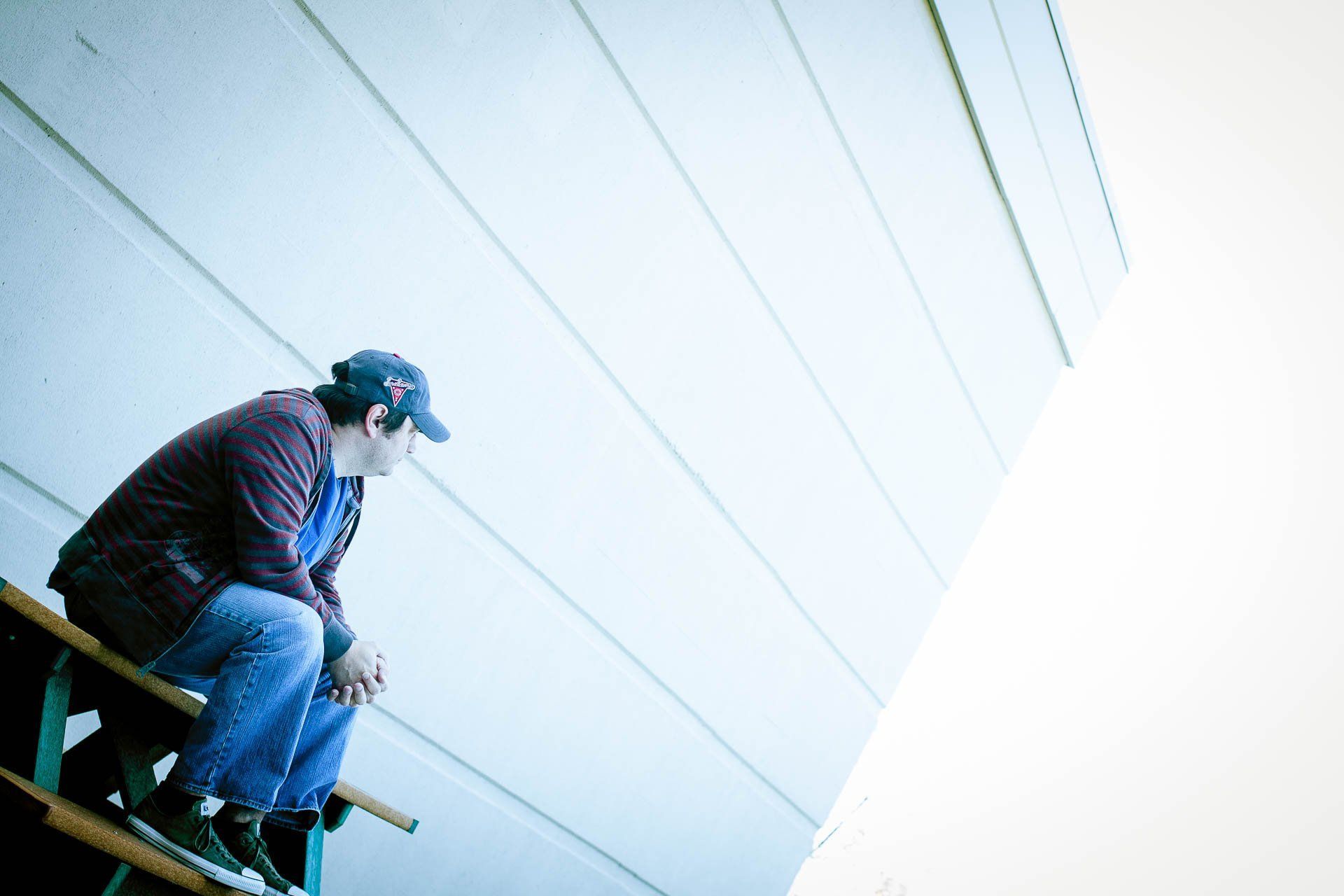 Person wearing a blue cap sits on a ledge beside a tall white wall, looking out.