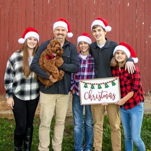 Family of five in Santa hats pose with a dog and a Christmas sign in front of a red barn in Carmel Indiana