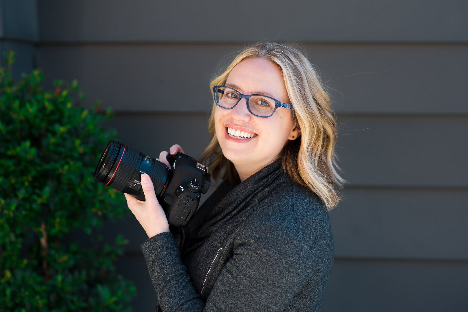 Photographer Kristin Madeja with glasses holding a camera outdoors beside greenery