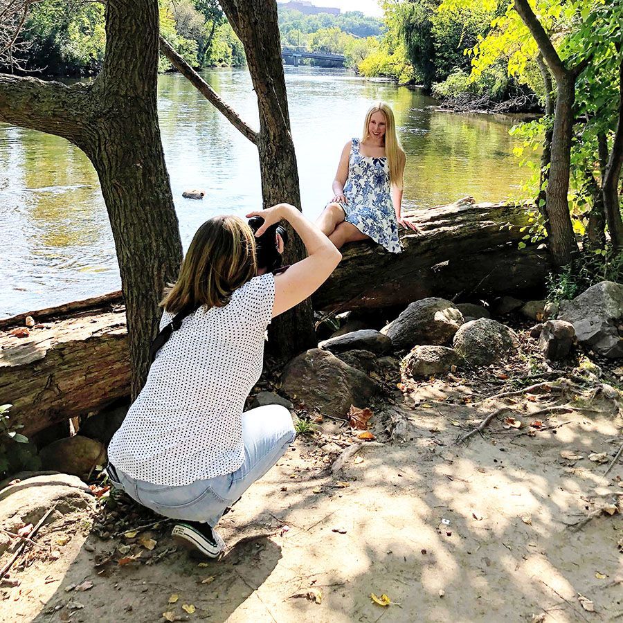 Photographer Kristin Madeja crouches by a river, taking a photo of a high school senior girl sitting on a log in a park.