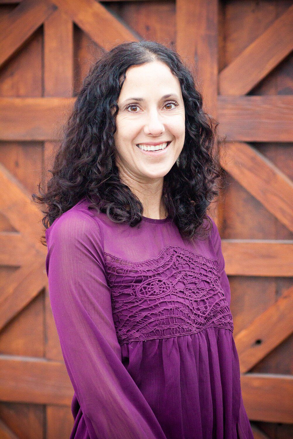 Headshot of an Fortville woman in a purple blouse smiling in front of a wooden gate