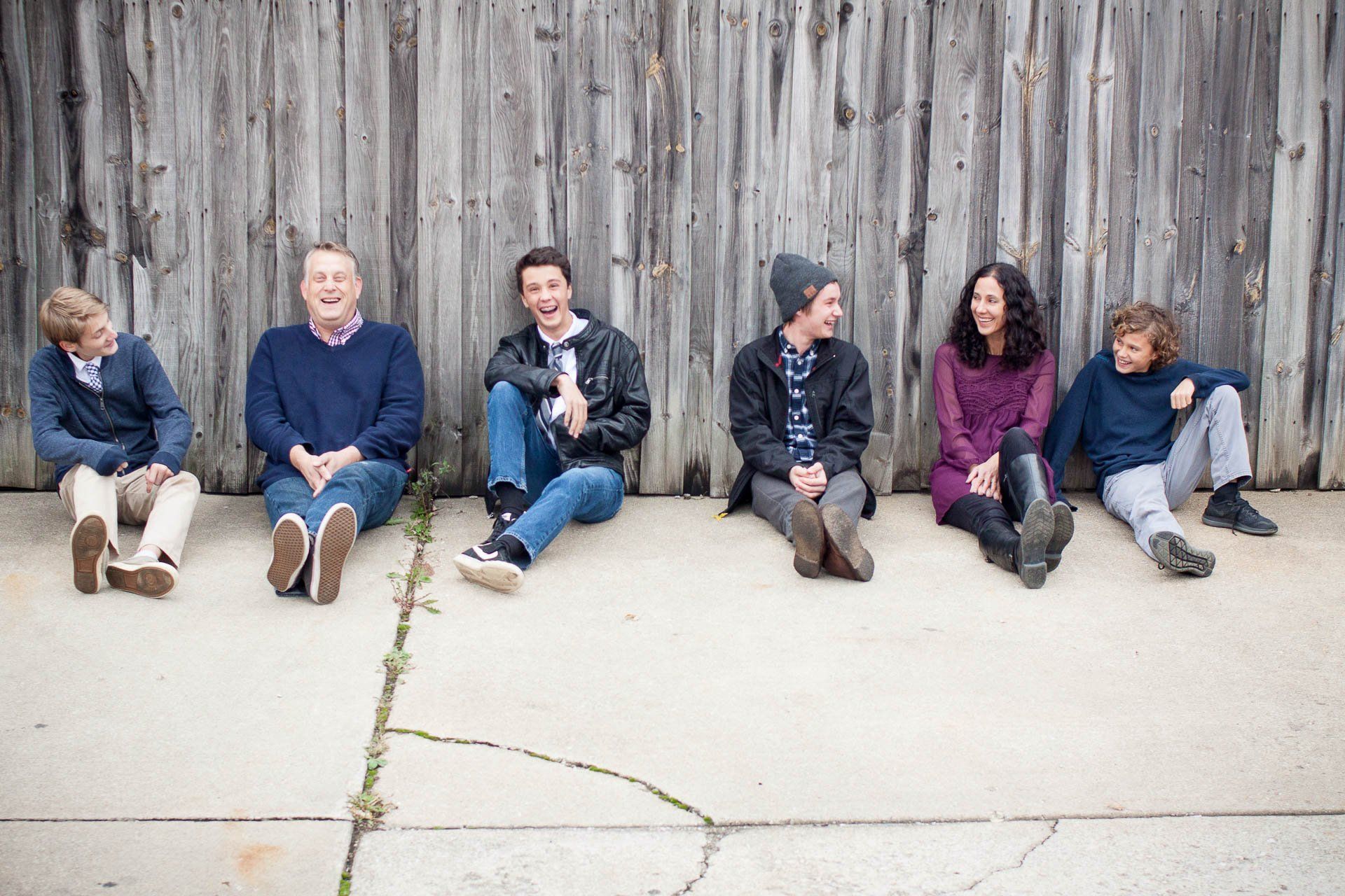 Six people sit against a weathered wooden fence on a concrete sidewalk in Pendleton, Indiana