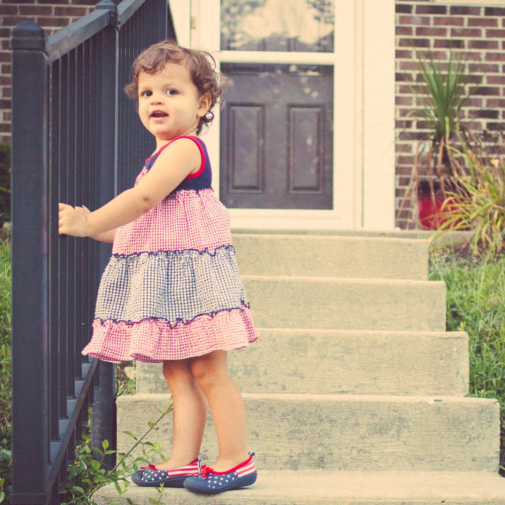 Toddler in a pink dress standing on outdoor steps by a black railing, looking back toward the camera in Fortville, Indiana