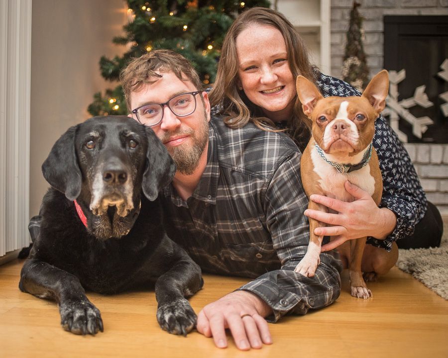 Two people posing with a black dog and a brown-and-white dog on a wooden floor indoors in Noblesville, IN.