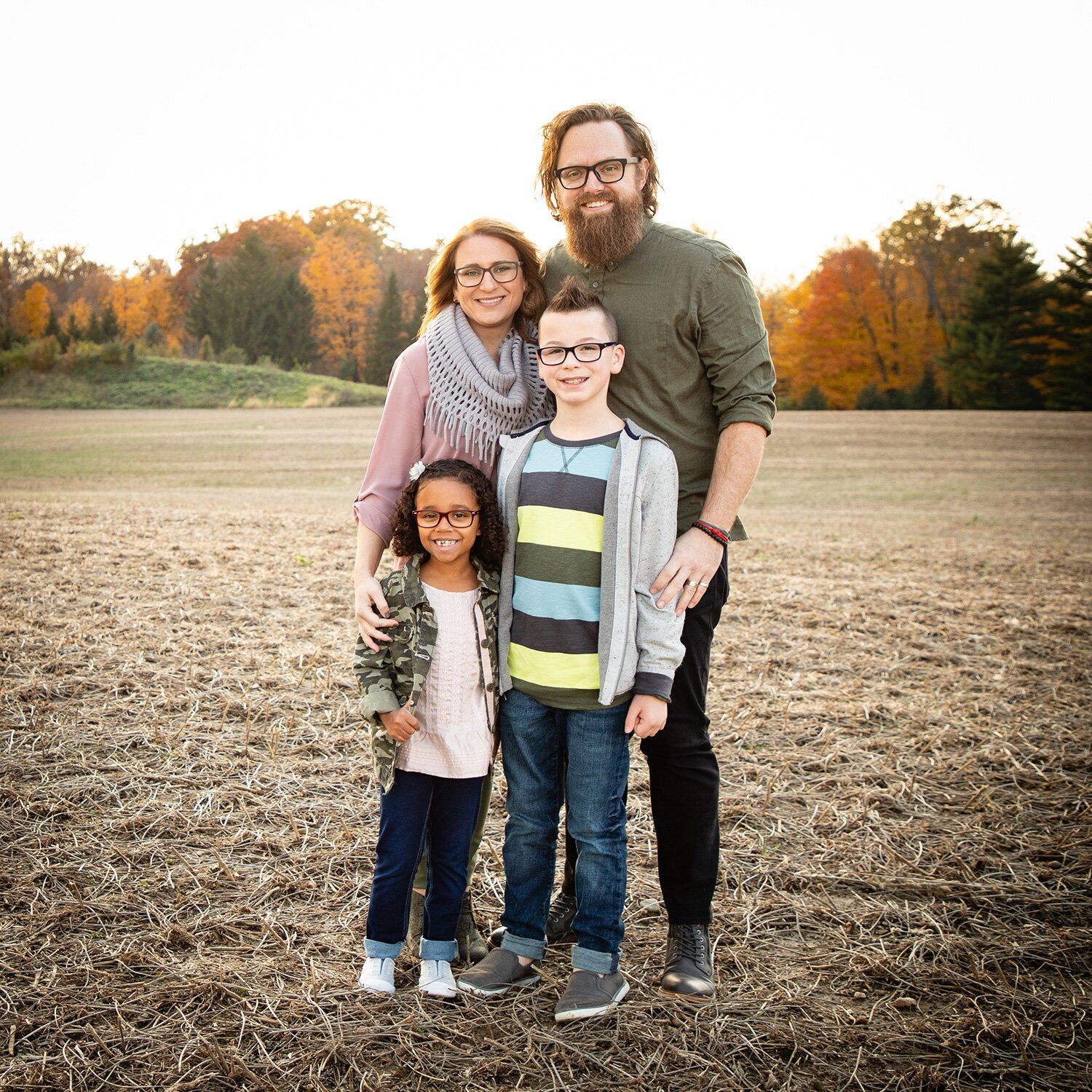 Family of four standing together in a field with autumn trees in the background in Noblesville, Indiana.